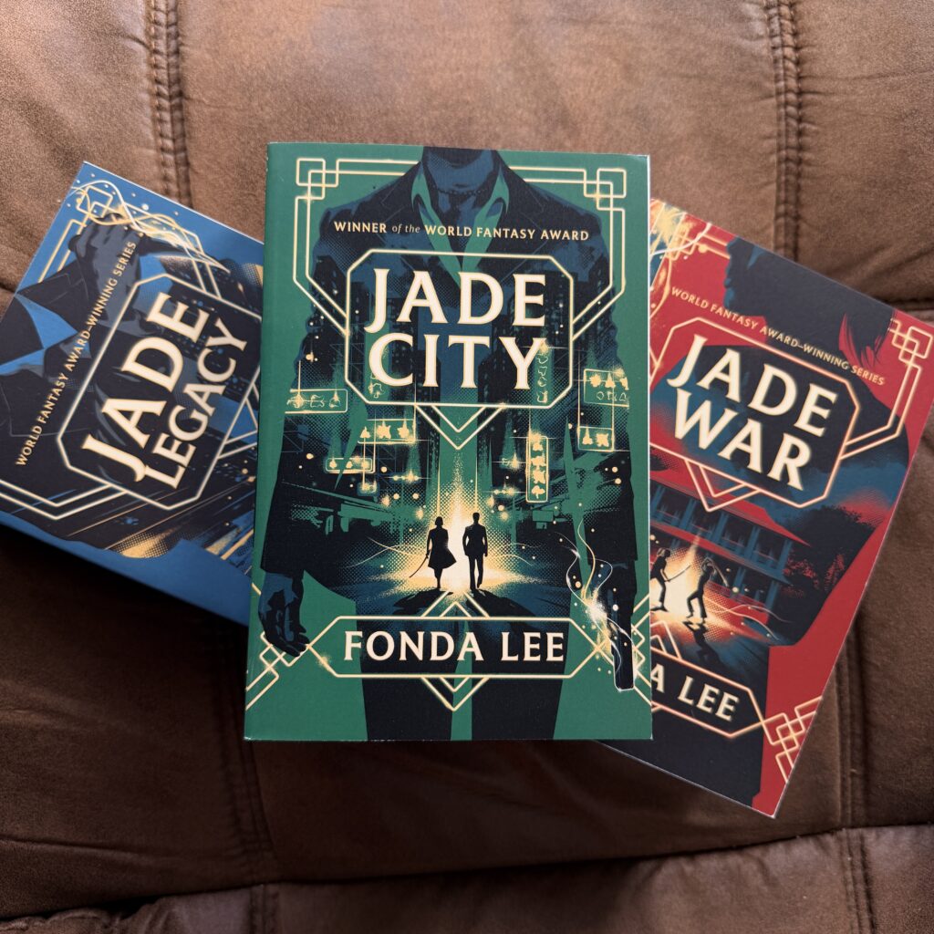 Jade City › 200 Fingers & Toes Three fantasy novels fanned on a brown couch, with the center green Jade City cover featuring geometric circuitry and silhouetted figures.