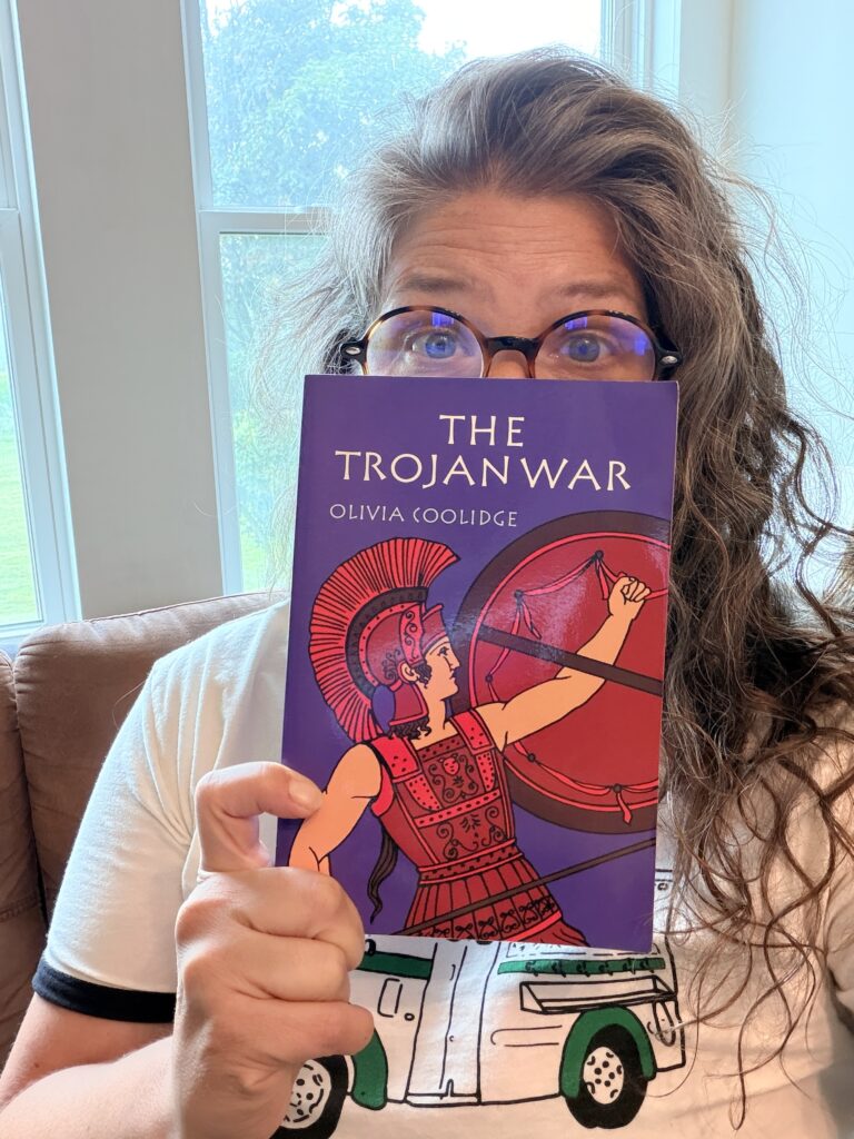 Person with round tortoiseshell glasses holding a purple book titled The Trojan War, covering their mouth.