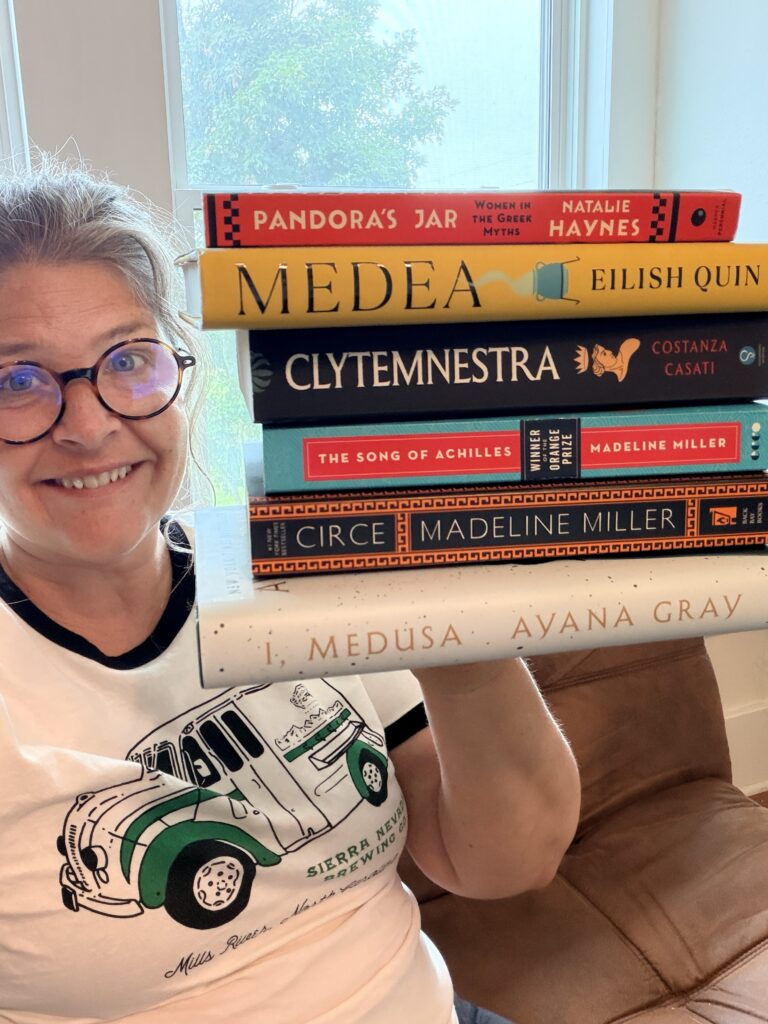 Amber holding a stack of Greek mythology books including Circe, The Song of Achilles, Clytemnestra, Medea, I Medusa, and Pandora’s Jar.